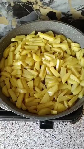 A close-up, top-down shot shows a frying pan filled with chopped potatoes. The potatoes are thinly sliced and appear to be seasoned with spices. They are arranged in a single layer across the pan. The pan is resting on a stovetop. The background is blurred, but suggests a kitchen environment.