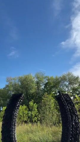 A video displays an outdoor scene featuring a sculpture made of black, interlocking materials, resembling two curved, portal-like structures. The sculpture is situated in a grassy area with trees and foliage in the background under a blue sky with some clouds. A person wearing a black t-shirt, brown pants, and a backpack walks towards the sculpture, stops in front of it, and turns to face the camera. The person appears to be posing for a photo. The lighting suggests it is daytime.