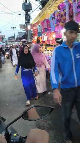 The video shows a busy street scene, likely a market, with stalls selling clothing. Several people are walking and riding motorcycles. In the foreground, a person is riding a motorcycle, with their hand visible on the handlebars. Further down the street, a woman in a blue dress and headscarf is walking, and behind her, another woman in a pink headscarf is walking. To the right, clothing racks are filled with colorful children's clothes. On the left side of the street, more people are walking, and there are motorcycles parked. The sky appears overcast, suggesting it might be early morning or late afternoon. The overall atmosphere is that of a bustling marketplace.
