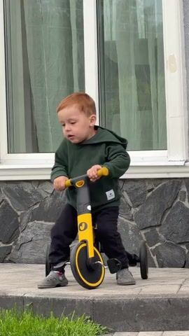 A young boy with red hair rides a yellow and black tricycle on a paved walkway. He is wearing a dark green hooded sweatshirt and black pants. The tricycle has three wheels, with two in the back and one in the front. The boy is looking to his right as he rides.

The scene appears to be outdoors, likely in a residential area. Behind the boy, there is a house with a stone foundation and large windows. A drainpipe is visible on the side of the house. There is also a black fence and some greenery in the background. The paved walkway is bordered by grass. The lighting suggests it is daytime, possibly overcast. No specific time of day or weather conditions are definitively indicated.

The tricycle has the word "Junior" visible on its frame. The boy's facial expression is neutral, and he seems to be focused on riding. The overall impression is of a child enjoying a simple outdoor activity. The location context provided is Tashtagol, Russia, which may inform the architectural style of the house and the type of fencing visible.