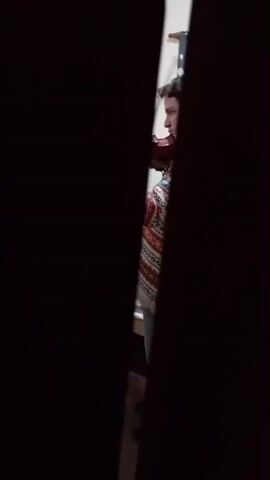 This media file shows a person playing a violin. The shot is taken from a side-angle, peering through a narrow gap in what appears to be a doorway or a crevice between two objects. The person, who is facing away from the camera, is wearing a patterned sweater with red, white, and black geometric designs. Their hair is dark and short. The violin is held in the typical playing position, under their chin, and the bow is in motion across the strings.

The background is dimly lit, with a hint of a white wall or surface visible behind the musician. A patterned blanket or rug is spread out on a surface below the person, suggesting they might be sitting on a bed or a couch. The lighting appears to be artificial, suggesting it could be daytime or nighttime, but the overall darkness of the scene makes it difficult to determine the time of day. The ambiance is intimate and private, as if the viewer is observing a moment of practice or personal expression from a concealed vantage point. No text is visible in the media file. The location context of Tolosa, Argentina, does not provide specific visual cues within the frame itself.