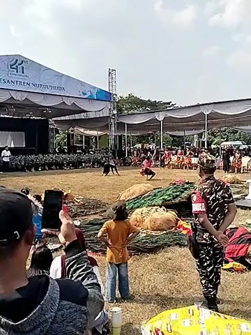 This is a video clip of an outdoor cultural performance. In the foreground, a person in a camouflage uniform stands facing away from the camera, facing the performance area. To the left, the backs of several people are visible, some holding up mobile phones to record. The performance area itself is a grassy field. In the mid-ground, two performers in traditional clothing are engaged in a dance or martial arts demonstration. One performer is on the ground, while the other is in mid-air, possibly leaping or performing a dynamic movement. They are surrounded by what appear to be decorative props, possibly related to a cultural art form like Barongan or Reog. In the background, a stage with a large tent structure is set up, with a banner displaying "24TH ANNIVERSARY PONDOK PESANTREN NURUL HUDA". A crowd of spectators is seated and standing behind the performance area, facing the stage. The sky is bright and appears to be daytime. The overall impression is of a community event or festival.