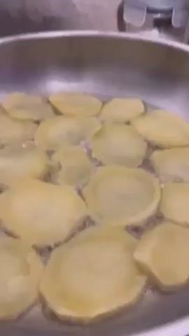 A close-up shot shows potato slices frying in oil in a silver-colored pan. The slices are mostly submerged and appear translucent. Tiny bubbles are visible around the potatoes. The background is out of focus, showing a countertop and a storage container with a blue lid.