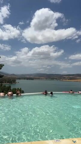 A group of people relax in an infinity pool with a scenic view of a lake and rolling hills under a blue sky with white clouds. Several individuals are visible in the water, some are sitting at the edge of the pool, and one person is lying on a pink float. The water in the pool is clear and inviting, and the sun is shining brightly. The surrounding landscape is dry and arid, with sparse vegetation. The scene conveys a sense of tranquility and relaxation.