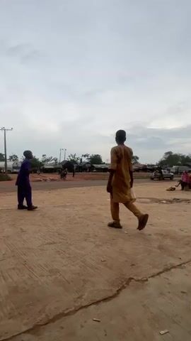 A vertical video displays a dusty outdoor scene, possibly a marketplace or roadside station, under an overcast sky. In the foreground, a person in traditional tan clothing walks away from the camera, carrying a light-colored bag. To the left, another person stands wearing a purple outfit. The background features several structures, including what appear to be gas station canopies with red and yellow accents. Various vehicles, including vans and motorcycles, are parked around the area. Several people are visible near the structures and vehicles, some sitting or standing. The ground is a mix of packed dirt and concrete. The overall impression is one of a busy, unpaved public space.