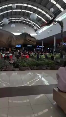 The video shows a large indoor space, possibly a mall or a food court, with a high ceiling featuring a grid-like structure and several large, white, spherical light fixtures. In the foreground, there is a large, realistic model of a Tyrannosaurus Rex dinosaur surrounded by artificial plants and rocks. Behind the T-Rex, there's an area with tables and chairs where people are sitting and presumably eating. Various food establishments can be seen in the background, including a Domino's Pizza outlet and what appears to be a Hudson store. In the upper right corner of the frame, there's a model of a Pterodactyl dinosaur suspended from the ceiling. The camera is positioned on a shiny, tiled floor, providing a view of the entire scene. The overall impression is that of a commercial space trying to attract customers with dinosaur-themed decor.