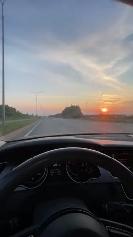 The video begins with a view from inside a car, looking out through the windshield. The car is on a highway, and the sun is setting in the distance, casting a warm glow across the sky. The dashboard of the car is visible, with a steering wheel and instrument cluster. The camera then pans to the right, showing a young woman sitting in the passenger seat. She is holding a smartphone and looking away from the camera. The video ends with a close-up of the woman's face.