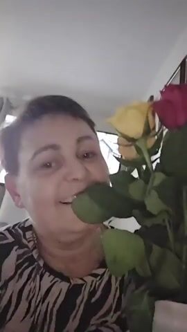 A woman with short brown hair and a patterned shirt smiles at the camera while holding a bouquet of yellow and pink roses. A television is visible in the background.