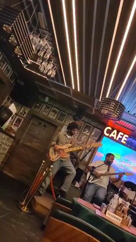 The video shows a band performing in what appears to be a cafe. Three men are visible on a raised stage. The man on the left is playing a light-colored electric guitar and wearing a light brown shirt and blue jeans. The man in the center is also playing a light-colored electric guitar and is wearing a white t-shirt and blue jeans. The man on the right is playing drums and is wearing a white shirt. Behind the band, a neon sign reads "CAFE 1986" with a screen displaying "US OPEN." The cafe has a wooden interior with modern lighting fixtures. The overall atmosphere suggests a lively indoor venue.