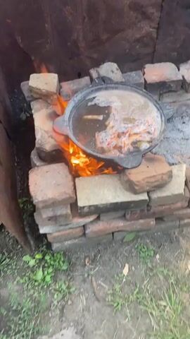 A close-up shot of a black pot filled with water and shrimp, cooking over an open fire. The pot is supported by a structure made of bricks. The fire beneath is bright orange and yellow, with flames licking the sides of the pot. The water is bubbling, and the shrimp are visible, some appearing pink and cooked. There are also some green leaves in the pot, possibly bay leaves. The background is blurred, but appears to be an outdoor setting with some greenery. The overall scene suggests a rustic, outdoor cooking experience.