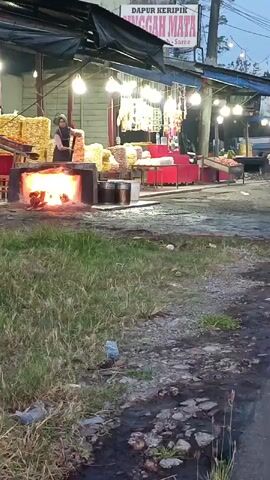 A street food vendor, wearing a dark hijab and light-colored shirt, is actively cooking at an outdoor stall. The stall is covered by a dark tarp and illuminated by strings of bright lights. In front of the vendor is an open fire pit containing bright flames, with metal pots nearby. The vendor is using a long-handled utensil, possibly a spatula, to stir or flip food over the fire. Stacks of what appear to be packaged snacks or ingredients are displayed behind the vendor and on red tables. The ground in front of the stall is a mix of dirt and patchy grass, with some debris visible. The overall atmosphere suggests an evening or dusk setting.