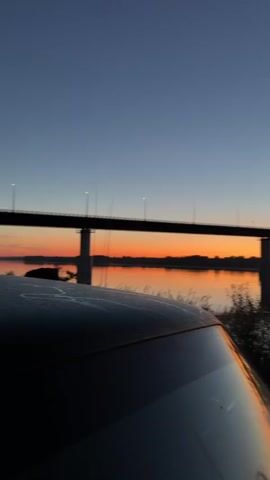 A vertical shot shows a bridge spanning across a body of water during sunset. The sky is a deep blue, transitioning to a vibrant orange and yellow horizon line above silhouetted land. The bridge's lights are visible, casting reflections on the water. In the foreground, the roof and windshield of a dark-colored vehicle are visible, with some foliage and grass on the right side. The water appears calm, with some ripples reflecting the colors of the sunset. The overall scene is peaceful and serene.