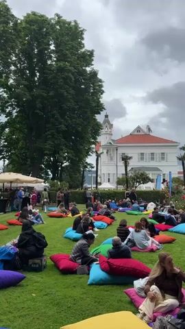 The video shows a group of people sitting on colorful beanbag chairs arranged on a grassy lawn. In the background, there is a large white building with a red roof and a clock tower. There are also trees, palm trees, and rose bushes visible. Some people appear to be interacting with each other, while others are looking towards the building. The sky is cloudy. The overall scene suggests an outdoor event or gathering.