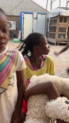 The video shows two young girls with braided hair in an outdoor setting. The girl in the foreground is wearing a white t-shirt with a rainbow star design and is looking down while manipulating something small in her hands, then puts it in her mouth. The girl behind her wears a yellow top and holds a tan teddy bear, looking toward the camera and smiling. In the background are other children, a shed, a chicken coop, and various other objects. The ground is dirt. The girls appear to be playing or interacting with each other.