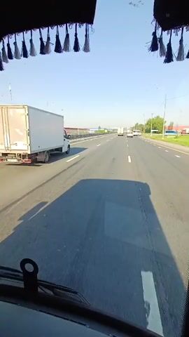 The camera, from inside a truck, looks out over a highway on a clear day. The road has multiple lanes and other trucks are visible in the distance. A building is visible to the left of the highway. Tassels hang from the top of the windshield.  The truck the camera is in casts a shadow on the road.