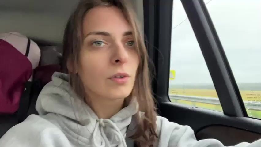 A young woman with brown hair and green eyes is in the passenger seat of a car. She is wearing a grey hooded sweatshirt. Bags are visible in the back seat of the car. The woman is looking at the camera and smiling. The weather outside appears overcast.