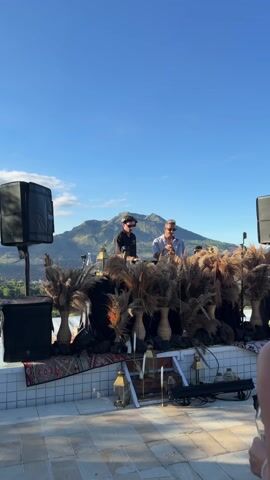 Two men, dressed in casual shirts and sunglasses, are DJing on an outdoor stage with a scenic mountain backdrop under a bright blue sky. The DJ booth is decorated with tall bouquets of pampas grass in rustic vases. A black cloth drapes across the front of the booth, adorned with small brass lanterns and candles. Speakers stand on either side of the booth. In the foreground, part of a patterned rug is visible on the tiled stage. One DJ is actively manipulating the equipment, while the other gestures as if talking to the crowd.