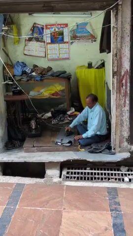 A man is seated on the floor of a small tailor shop, working on a piece of gray fabric. He is wearing a light blue collared shirt and dark pants. In front of him is a sewing machine, various pieces of fabric, and other sewing supplies. The shop is cluttered with items, including clothes hanging from the wall and a yellow cloth draped over a shelf. There are calendars on the wall and a glass display case filled with fabric. The shop appears to be open to the street, with a metal grate in the foreground.