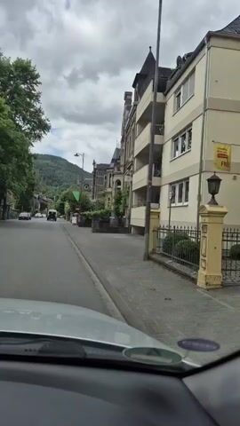 The video shows a first-person perspective from inside a moving vehicle, traveling down a street lined with buildings and trees. On the right, there are multi-story buildings, some with ornate architecture and balconies. On the left, there are also buildings, some partially obscured by trees. A tall green tree with scaffolding around it is visible further down the street on the left. In the distance, hills or mountains covered in greenery can be seen under a cloudy sky. Several cars are parked along the street or driving in the same direction. The vehicle appears to be moving at a slow to moderate speed.