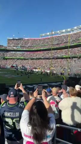 The video shows a packed stadium, with a football field in the center and spectators filling all the seats. Green and white smoke billows into the clear blue sky. Many spectators are holding up their phones to capture the scene. Some are wearing jerseys, including one with "CHANCELLOR" and "31" on the back. People are clapping and cheering. The overall atmosphere appears to be one of excitement and celebration.