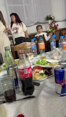 The video shows a cluttered table at what appears to be a wedding reception or similar celebration. The table is covered with a white tablecloth and is littered with half-eaten plates of food, empty or partially empty bottles of water, Coca-Cola, and Red Bull. An empty liquor bottle is visible towards the back. A child is visible sitting at the table in the background, while an adult stands nearby. A man with a beard can be seen sitting at another table. The room is brightly lit and decorated with flowers.