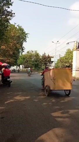 A street scene with several motorcycles and a cart. On the left side of the road, a motorcycle is parked by a stall with two red umbrellas. A person in a red shirt is sitting on the motorcycle. Further down the road, a cart with a large tan box is being pulled by a motorcycle. Several other motorcycles are visible further in the distance. The road is paved and lined with trees. The sky is clear and blue, indicating it is daytime.