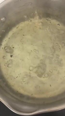 The video shows a close-up, top-down view of a pot of boiling water. The water is clear with small bubbles actively rising to the surface. Some darker particles are visible suspended in the water, possibly food remnants or impurities. The pot itself is made of brushed stainless steel and has measurement markings on the inside, indicating 2.5L and possibly another unmarked line. The pot is sitting on a dark surface, likely a stovetop. The lighting appears to be indoor ambient light, suggesting it could be any time of day. There are no people or animals visible in the frame, and no distinct location cues beyond what can be inferred from the object in the frame. The activity is simply the boiling of water, a common preparation step for cooking. There are no strong emotional cues conveyed by the image.