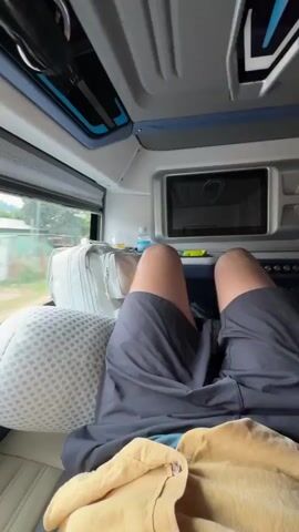The camera is inside a vehicle, likely a bus, moving along a road. A person is seen relaxing on a seat, with legs visible and supported by a pillow. The view outside the window shows a landscape of buildings, trees, and hills under a cloudy sky. The road is marked with white lines.