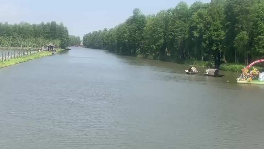 The video shows a wide river flowing horizontally from left to right. Both sides of the river are lined with tall green trees. On the left side, a small gazebo-like structure is visible near the edge. A small bridge can be seen in the distance on the left. In the river, there are two small, dark-colored boats. Further to the right, there's a floating platform decorated with a red archway, flowers, and what appears to be a waterfall feature. The day is bright and sunny. People's voices are heard in the background.