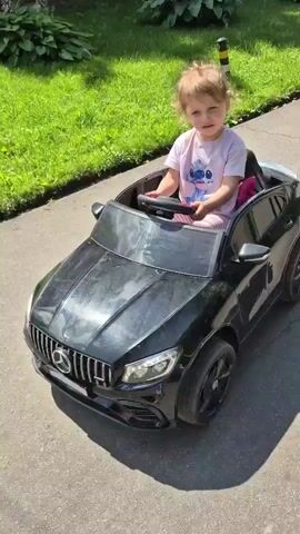 A young child sits in a black toy car, appearing to be a Mercedes-Benz model, on a paved surface. The child is wearing a pink t-shirt and has blonde hair. They are holding onto the steering wheel. The toy car is black with a prominent front grille and headlights. The background shows a grassy area with some plants and a building. The lighting suggests it is daytime.