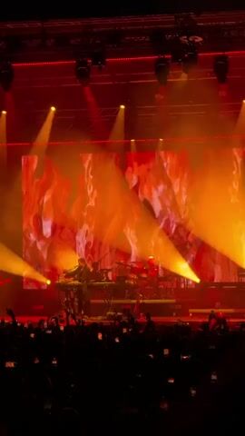 A concert stage with a large red backdrop featuring animated graphics. A male performer stands center stage, playing a keyboard. Behind him, a drummer is visible behind a drum kit. Red stage lights are set up along the front of the stage, and spotlights shine down from above. A crowd of people is visible in the foreground, many holding up their phones to record the performance. The overall atmosphere is dark and energetic, with the red lighting creating a dramatic effect.