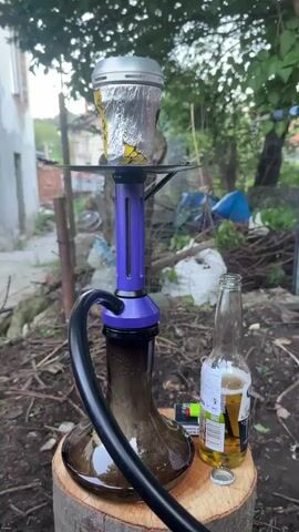 The video shows a hookah set up outside, next to a bottle of Corona beer. The hookah is primarily purple, black, and silver. The hookah bowl is covered in aluminum foil. The hookah sits on top of a wooden stump. Behind the stump is a cluttered yard with a building in the background. A lighter is on the stump next to the beer. There are sounds of someone smoking and other voices can be heard.