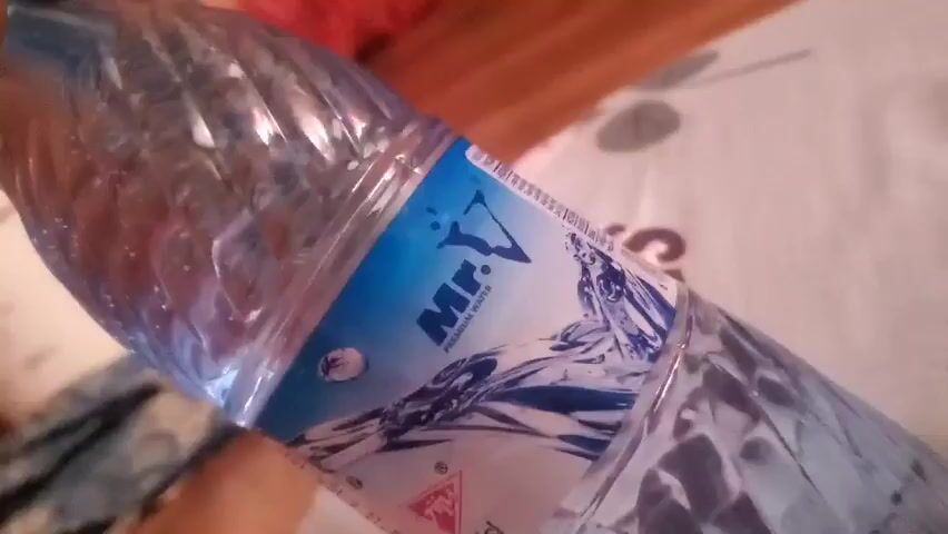 The video shows a close-up of a plastic bottle of water labeled "Mr. V PREMIUM WATER". The bottle is clear and filled with water. The label is blue and white with an image of water splashing and a drawing of a person. The bottle is resting on a patterned surface, possibly a tablecloth or bedspread. The background is slightly out of focus and appears to be a wooden surface and a patterned fabric. The lighting is dim.