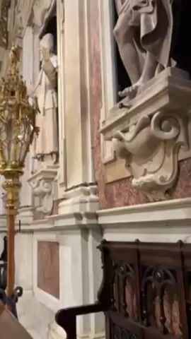 The video shows the interior of a church with ornate architecture. The walls are adorned with marble paneling and sculpted figures. Golden candelabras stand on either side, and stained glass windows provide illumination. The camera pans upwards, revealing a detailed ceiling and a structure supported by marble columns, above the altar. The floor is covered with a patterned tile design. The overall aesthetic is one of elaborate religious art.