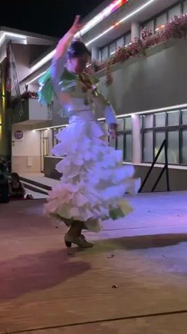 A woman in a white flamenco dress with ruffles dances on a wooden stage under purple and pink lights. She has her hair up with flowers and is wearing a white jacket with green accents. Her arms are raised and her body is angled to the right. In the background, a building with windows and lights is visible, and a few people are sitting on the ground to the left.