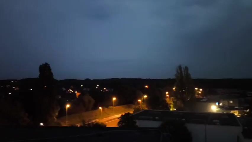 The video depicts a nighttime cityscape under a cloudy, dark blue sky. Streetlights and the occasional lit window illuminate buildings and trees. The scene is somewhat grainy, suggesting low-light conditions and possibly a mobile phone camera. No people are visible.