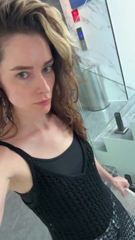A woman with wavy brown hair is walking down a modern marble staircase. She is wearing a black tank top under a black crocheted cover-up and black flip-flops. The staircase has glass railings and metal posts. An elevator is visible in the background. She looks at the camera briefly and then continues down the stairs.