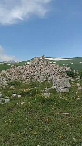 A rocky mound made of piled stones is in the center of the frame. The mound is large and uneven, with smaller piles of stones scattered around it. The area is a grassy field with patches of yellow wildflowers. In the background, there are green hills with snow on them, and a blue sky with white clouds. The camera is panning from left to right, showing the scene in a wide view.