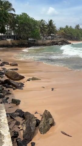 The video shows a tropical beach with light brown sand and dark rocks. Waves are breaking on the shore, and the water is a mix of turquoise and white. In the background, there are palm trees and buildings. The sky is blue with some clouds. No people are visible in the video.