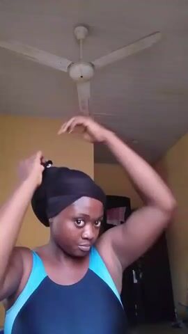 A dark-skinned woman with a black head wrap is adjusting her swimsuit. She is wearing a blue and black swimsuit and her hair is covered. Her arms are raised as she adjusts the wrap. The background is a yellow wall and a white ceiling fan.