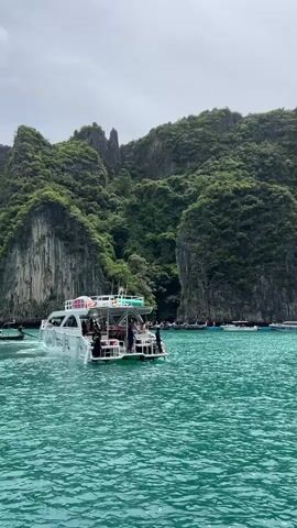 A white tourist boat with the logo "WAVA TRAVEL" is moving through turquoise water. Several people are visible on the back deck of the boat. In the background, there are steep, lush green cliffs that rise from the water. The sky is overcast.