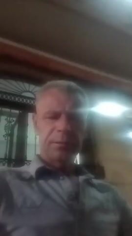 A man with short gray hair, wearing a gray collared shirt, is filmed from the chest up. He is looking directly at the camera with a neutral expression. The background is blurry and dimly lit, suggesting an outdoor setting at night, possibly a cafe or restaurant with patrons and lights visible in the distance. The lighting is artificial and casts a glow on the man's face.