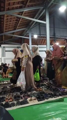 The video shows a group of women, most of whom are wearing hijabs, standing in a line and holding small boxes. They appear to be inside a large, open-sided building with a wooden ceiling and exposed metal beams. Shoes are neatly arranged on a green tarp in the foreground. Some of the women are reaching out to take items from the boxes, while others are looking down at them. The lighting is somewhat dim, with a bright light visible near the ceiling. The overall atmosphere seems to be one of a gathering or event, possibly a religious or cultural ceremony.