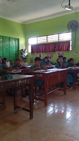 The video shows the interior of a classroom with approximately ten children, mostly boys, seated at wooden desks and appearing to be engaged in a lesson. They are all wearing green shirts and blue pants or skirts, indicating a school uniform. The room has green walls, tiled floors, and large windows with blinds and red curtains. A ceiling fan is visible on the wall, along with a clock and framed pictures. There is a projector mounted on the ceiling, and a cabinet with glass doors stands against the back wall, containing various items. The overall atmosphere suggests a typical school setting during a class.