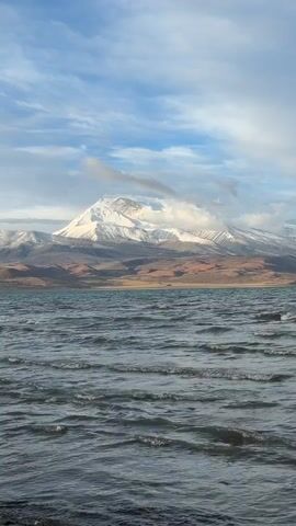 The video shows a wide shot of a lake with snow-capped mountains in the background under a partly cloudy sky. The water is choppy, with small waves visible across the surface. The mountains are partially covered with snow and surrounded by lower, brown hills. Clouds partially obscure the peak of the highest mountain. The sky is blue with patches of white and grey clouds.