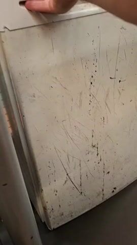 The video begins with a close-up of a scratched and dirty white refrigerator door. A hand is visible reaching to open it. The door swings inward, revealing the interior of the refrigerator. The refrigerator is sparsely stocked with a few items, including two bottles of water and a small dark bottle on the bottom shelf. The interior shelves and walls of the refrigerator appear somewhat grimy. The lighting inside the refrigerator is a warm yellow. The video then shows the door being closed again.