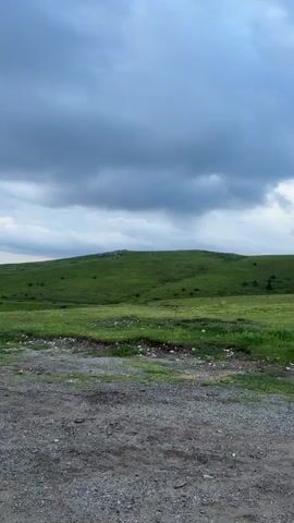 The video shows a scenic view of rolling green hills under a cloudy sky. The camera pans from left to right, revealing a gravelly road in the foreground and a vast expanse of green landscape stretching into the distance. In the background, layers of mountains are visible, with the sky dominated by heavy, dark clouds interspersed with patches of lighter sky. The overall impression is one of a dramatic, natural landscape, possibly during or just before a rain shower. There are no people or man-made structures prominently featured in the video, emphasizing the wild and open nature of the environment.