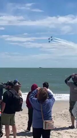 Here is a concise description of the video: 

The video shows a crowd of people standing on a beach watching a group of four fighter jets fly in formation overhead. The sky is blue with some clouds, and the jets leave trails of white smoke behind them. The beachgoers are facing the water, with some taking photos or videos of the spectacle. In the distance, a couple of boats can be seen on the water.