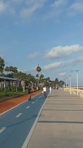 The video captures a scenic promenade along the coast, likely in Yenişehir, Turkey. The time of day appears to be late afternoon or early evening, with the sun casting long shadows across the paved walkway. The weather is pleasant, with a clear blue sky dotted with fluffy white clouds.

The promenade is divided into distinct paths: a vibrant orange jogging track runs alongside a bright blue cycling path, separated by a white line. A wide concrete walkway, presumably for pedestrians, stretches further along the coastline. Lining the promenade is a lush row of palm trees, creating a tropical ambiance. Beyond the trees, glimpses of buildings and structures can be seen, suggesting a developed urban area adjacent to the coast. A prominent flag pole with a red flag is visible in the distance, adding a nationalistic touch to the scene.

Several people are enjoying the recreational facilities. A person is seen cycling on the blue path, while another individual is riding what appears to be a scooter or electric bike on the same path. Further down the walkway, two figures are walking, one in white attire. A few other individuals are scattered along the promenade, engaged in walking and cycling, indicating a relaxed and active atmosphere. The presence of lampposts along the pedestrian path suggests that the promenade is also used in the evening.

The overall mood is one of tranquility and leisure, with people taking advantage of the beautiful coastal scenery for outdoor activities. The combination of the natural elements—sea, sky, and palm trees—with the well-maintained recreational infrastructure creates an inviting public space. The scene is peaceful and depicts a typical day at a popular seaside location.