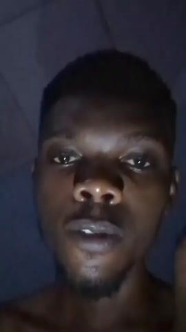 A dark-skinned man with short hair and a goatee is looking directly at the camera. He has a piece of food in his hand and puts it in his mouth. The background is dark and somewhat obscured.